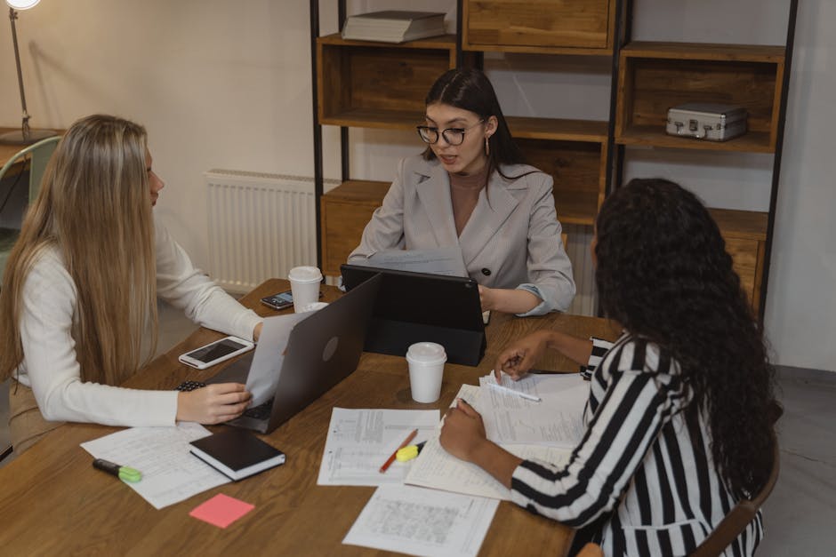 Three women engaged in a business meeting around a wooden table, sharing ideas and using digital devices.